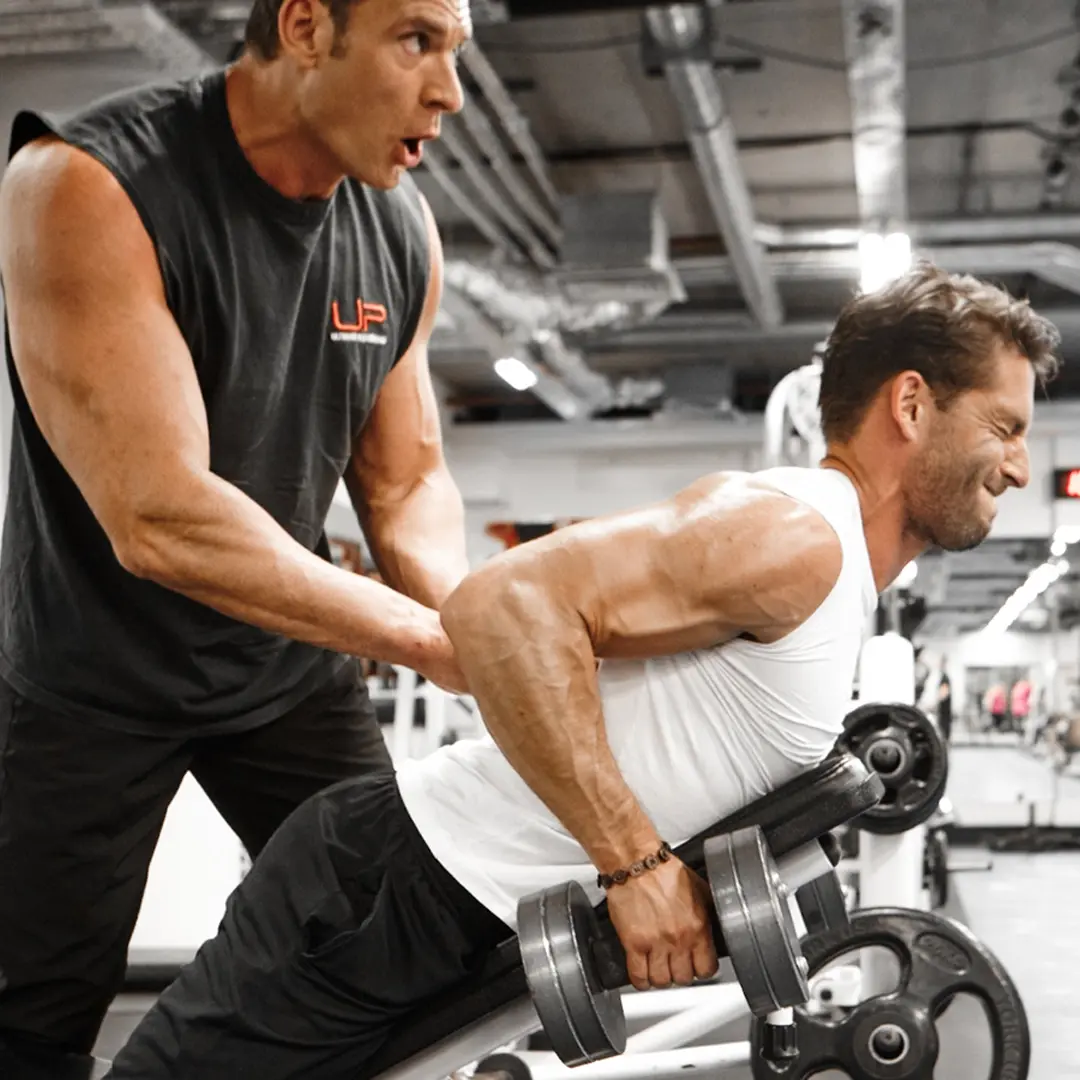 Man in white tank top lifting weights on an incline bench while another man in a sleeveless shirt spots him in a gym.