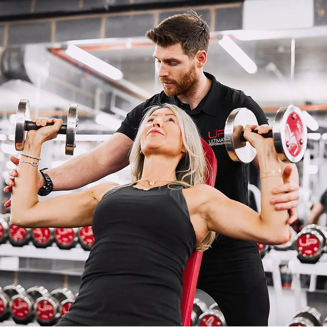 Female client lifting dumbbells during shoulder workout with male personal trainer's support in gym.