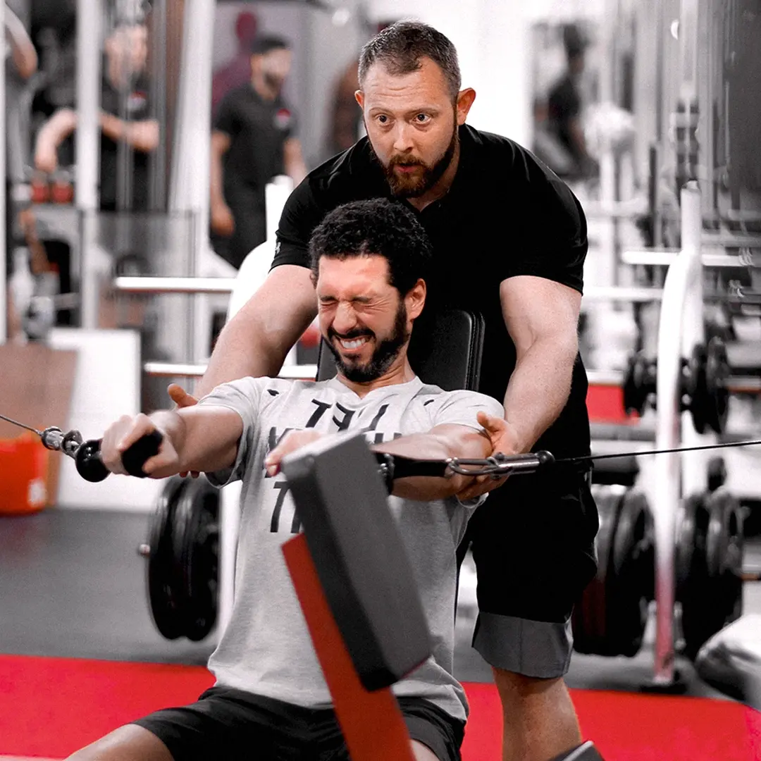 Fitness trainer spotting a man performing seated cable rows in a gym.