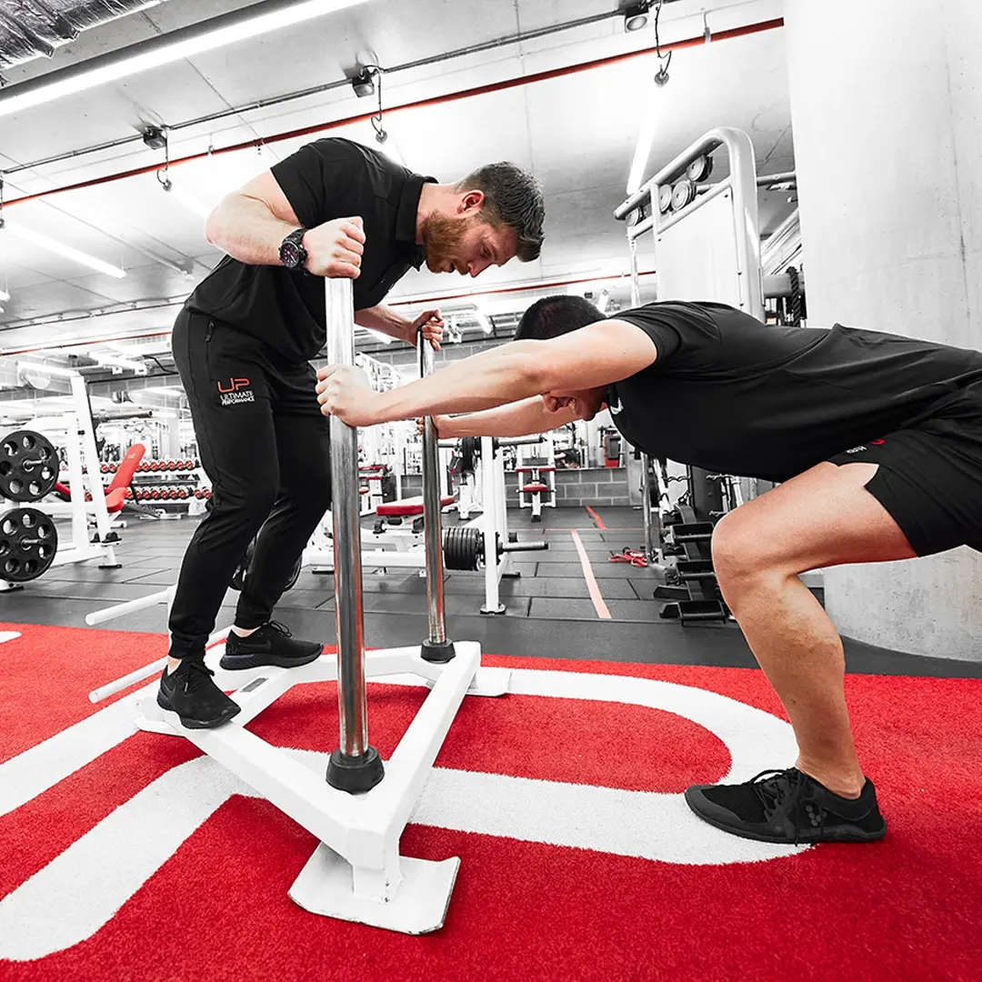 Personal trainer assisting man performing sled push exercise in gym with red floor mat.