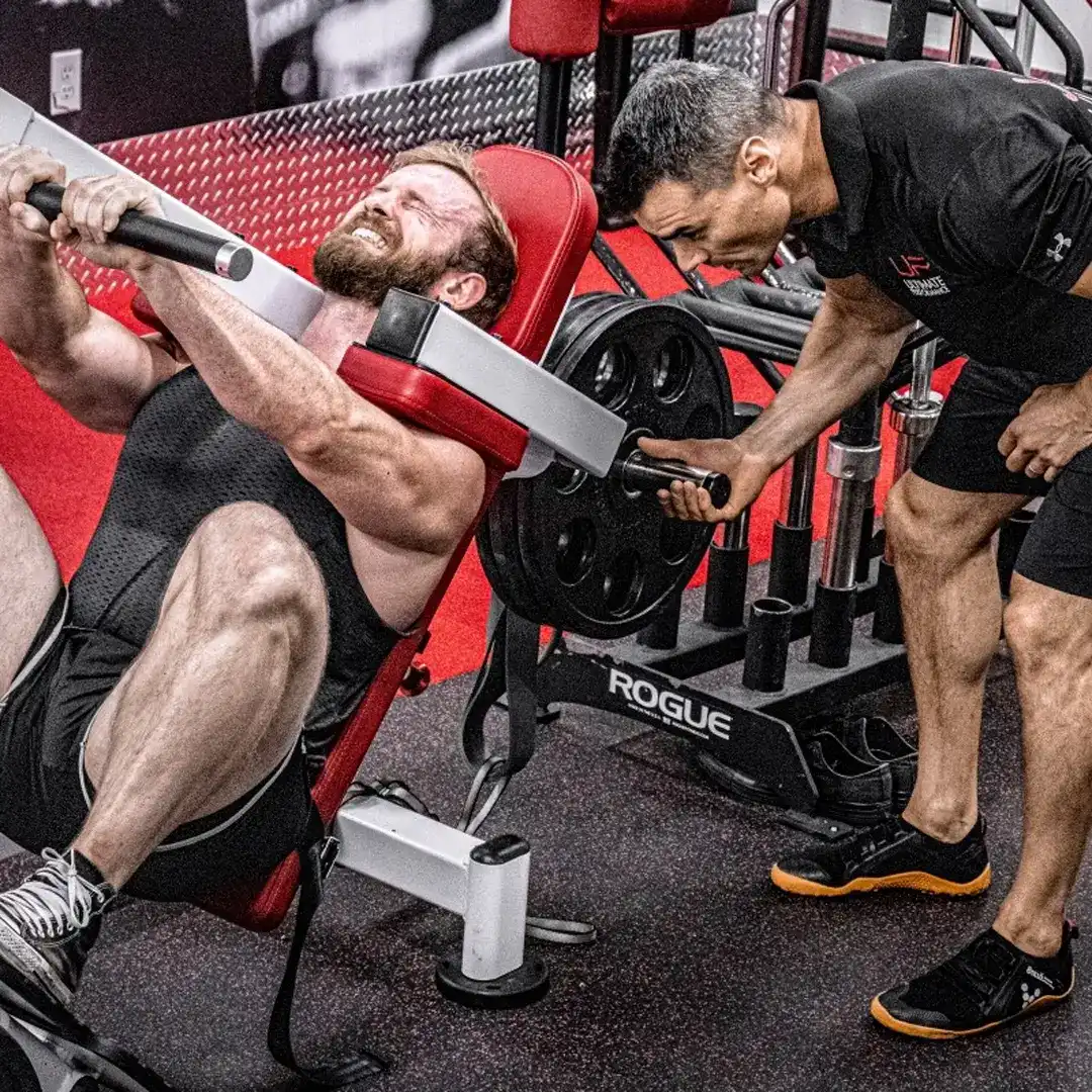 Man struggling with weight on a leg press machine while a trainer spots him in a gym.