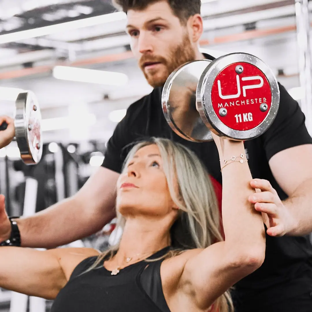 Woman lifting 11 kg dumbbells while a man spotter assists her in a gym.