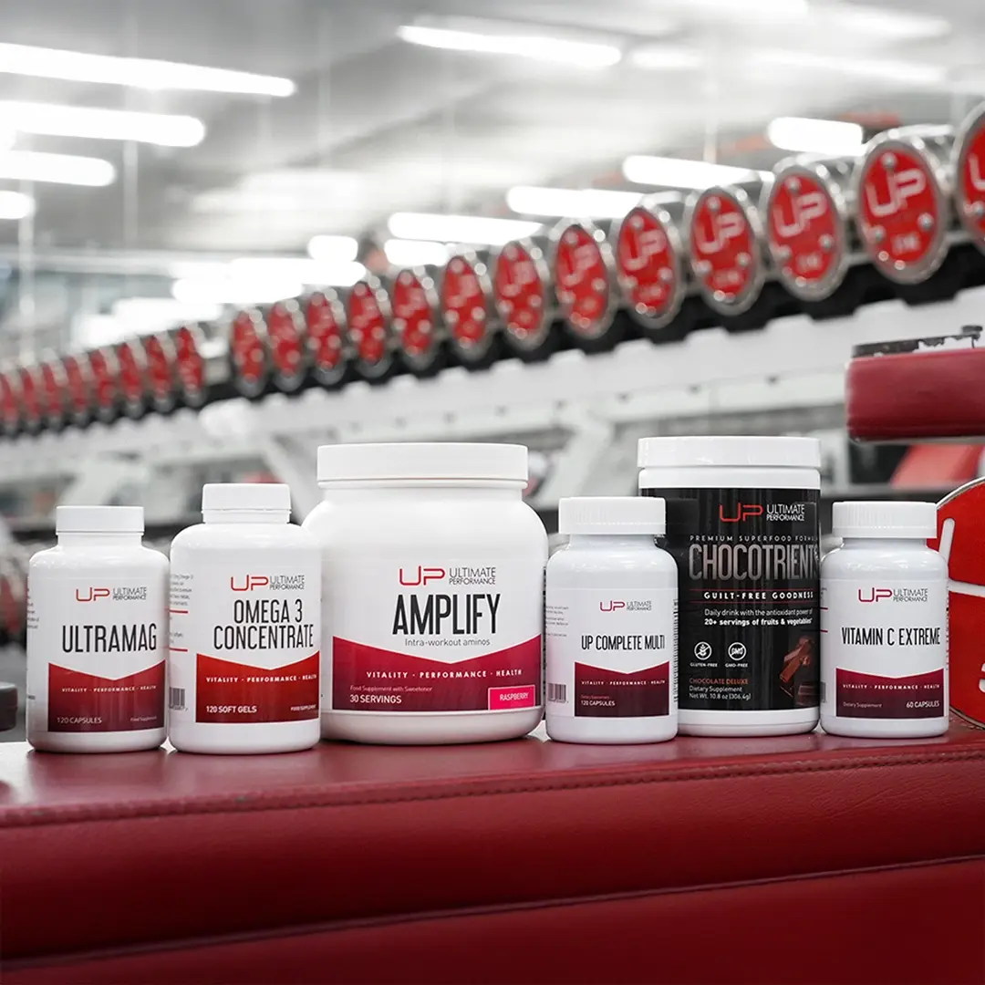 Six Ultimate Performance supplement containers including Ultramag, Omega 3 Concentrate, Amplify, UP Complete Multi, Chocotrient chocolate superfood, and Vitamin C Extreme on a red gym bench with dumbbells reflected in the mirror behind.