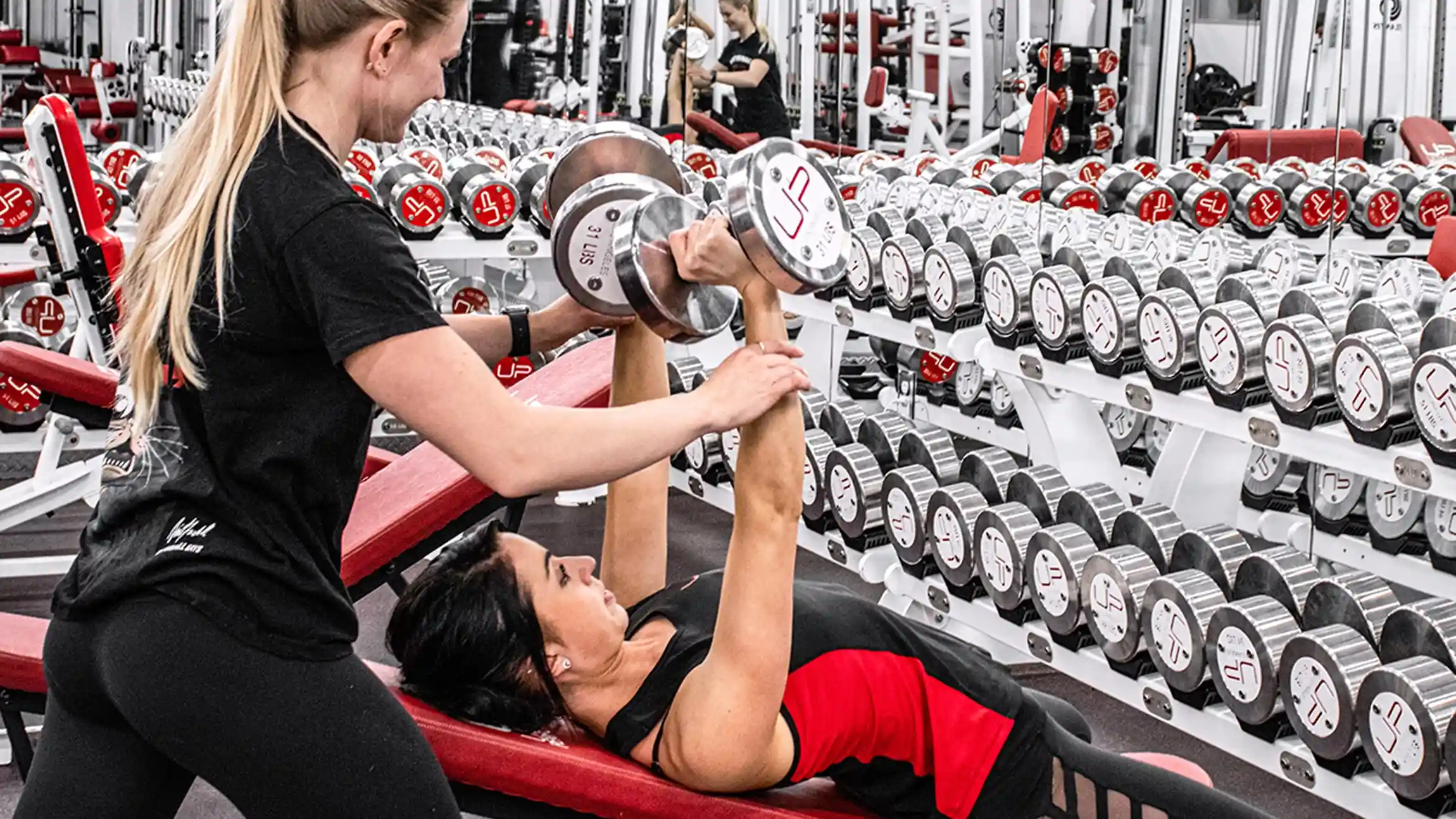 A woman lies on a red bench lifting dumbbells while a trainer assists her in a gym with rows of dumbbells on racks behind them.