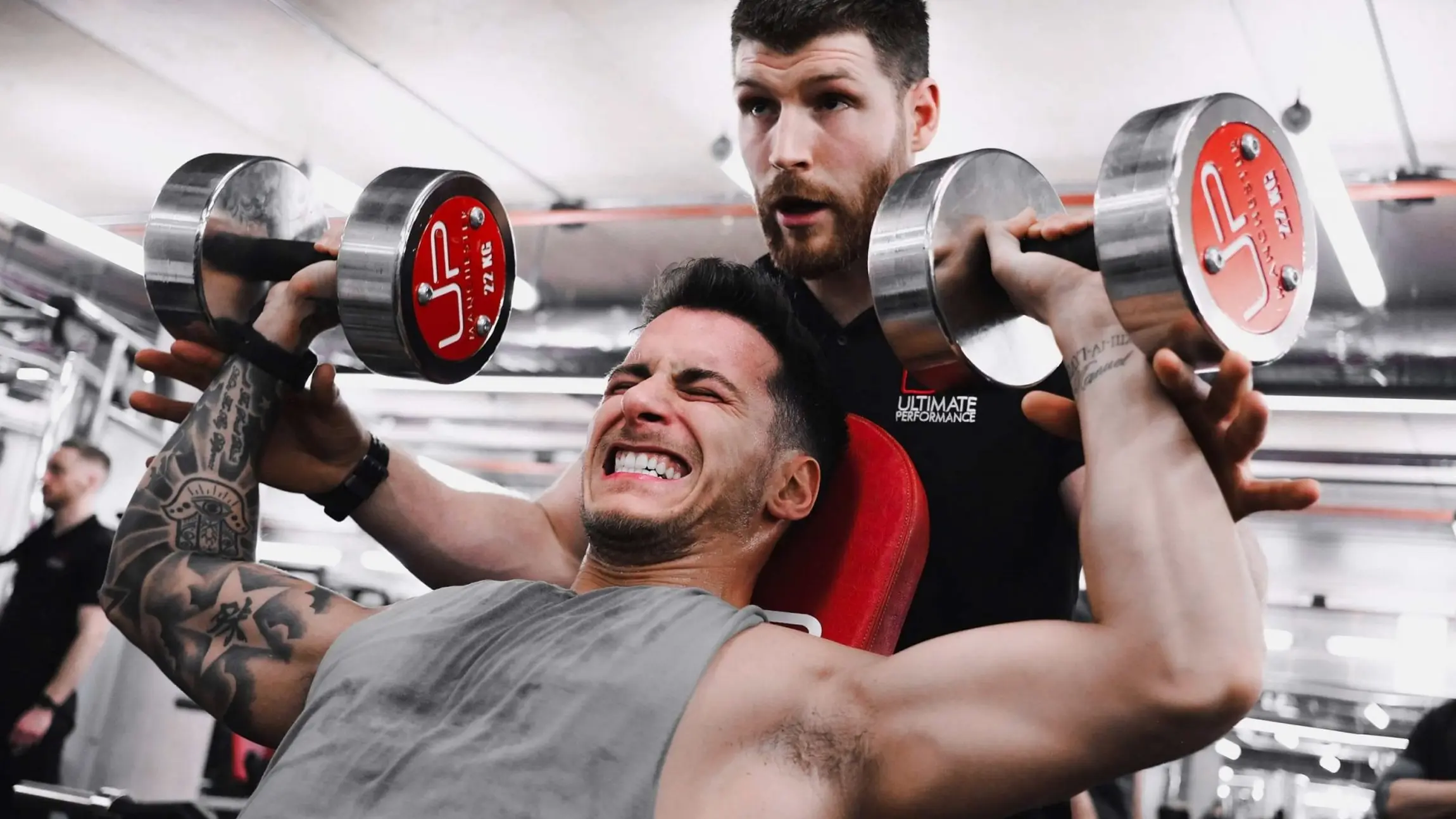 Man lifting dumbbells on a bench press with a trainer spotting him in a gym.