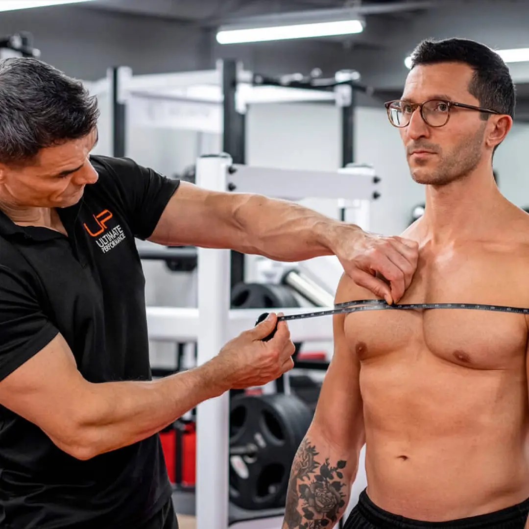 Trainer measuring the chest circumference of a shirtless man with glasses in a gym.