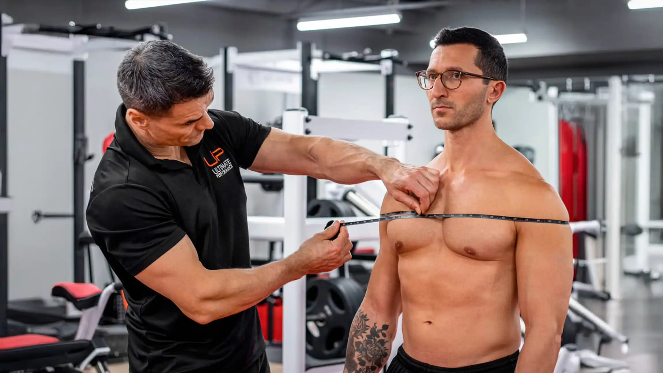 Trainer measuring chest circumference of shirtless man with glasses in a gym.