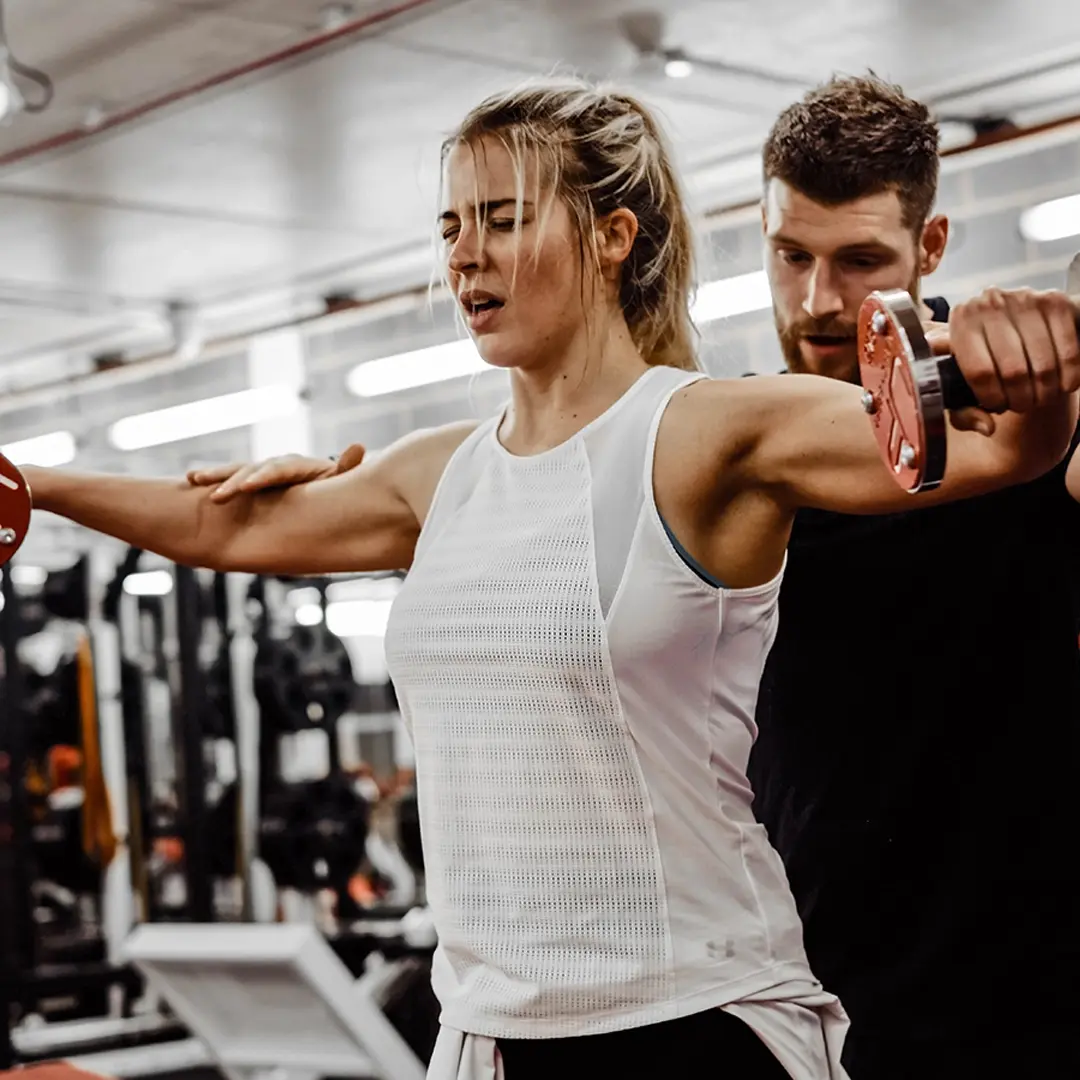 Woman lifting dumbbells with personal trainer spotting her in a gym.