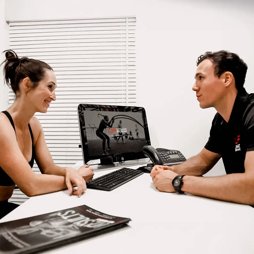 Two people facing each other in an office with a computer showing a workout scene and a magazine on the table.