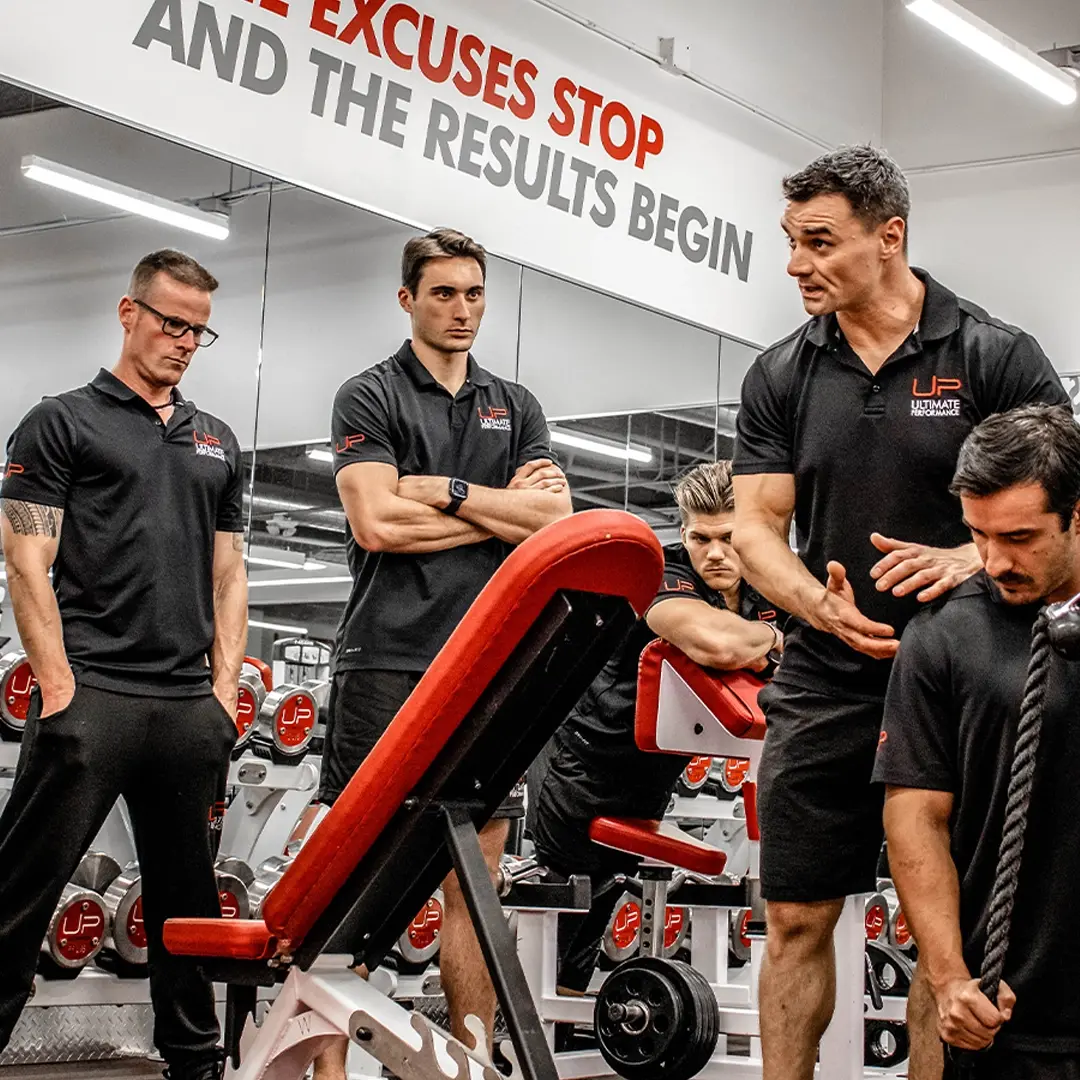 Four men in black Ultimate Performance shirts in a gym with a motivational sign reading 'NO EXCUSES STOP AND THE RESULTS BEGIN' behind them.