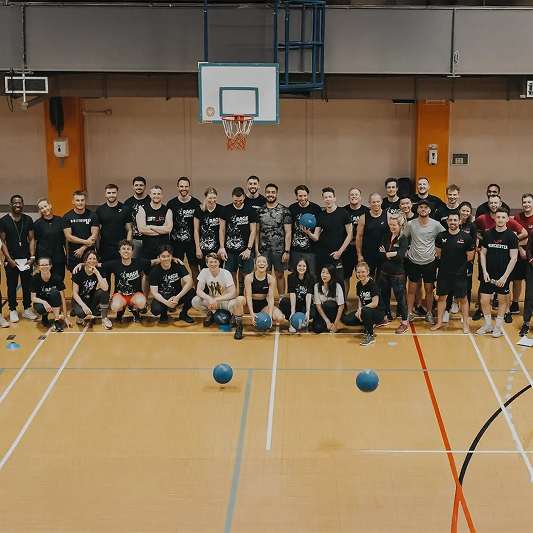 Large group of people posing in a gymnasium with blue dodgeballs on the floor and a basketball hoop overhead.