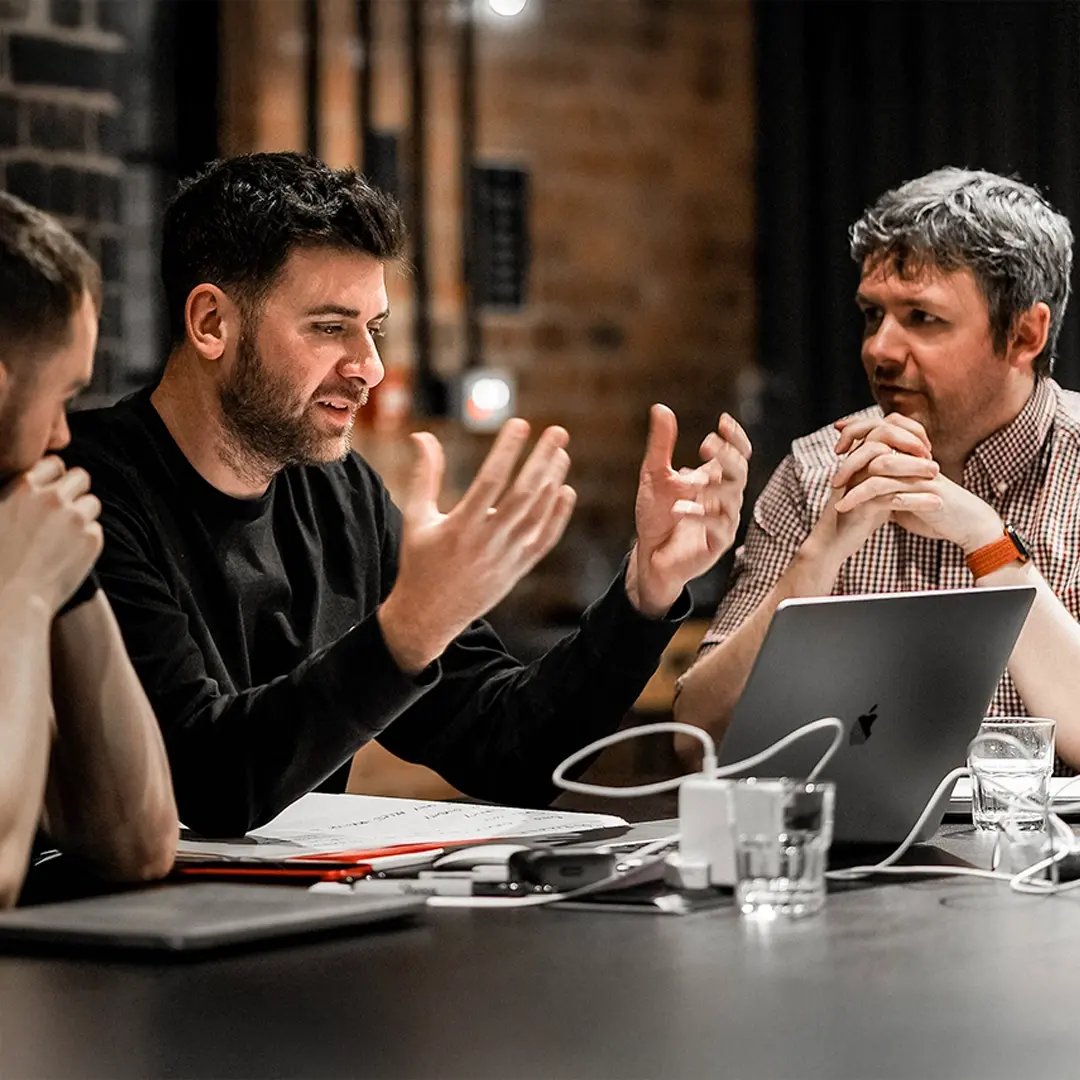 Three men in a business meeting with a laptop and documents, one man speaking with hands raised.