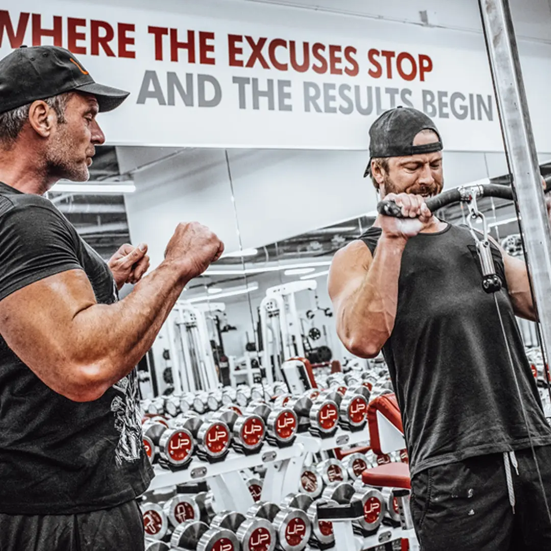 Man in black cap doing lat pulldown exercise under the guidance of a trainer in a gym with dumbbells and motivational sign in background.