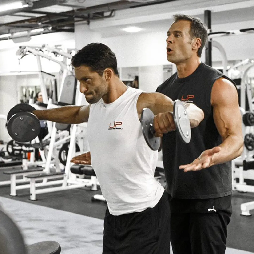 Man lifting dumbbells performing a lateral raise guided by a muscular personal trainer in a gym.