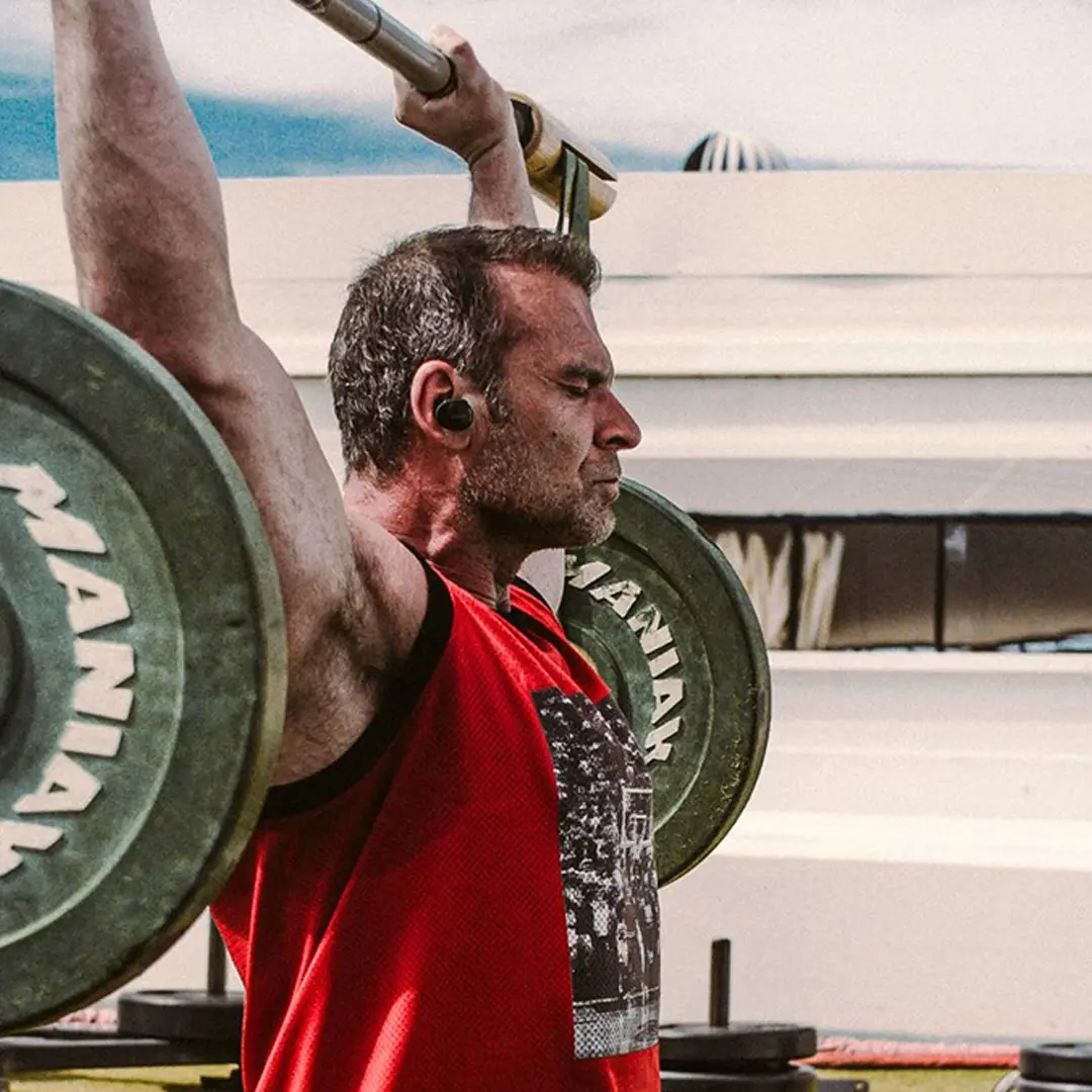 Man wearing a red tank top lifting a barbell overhead during an outdoor workout.