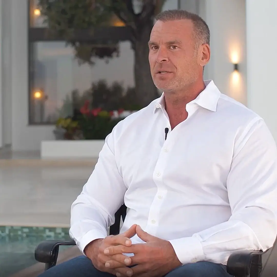 Man wearing a white shirt sitting outdoors, speaking in an interview setting.