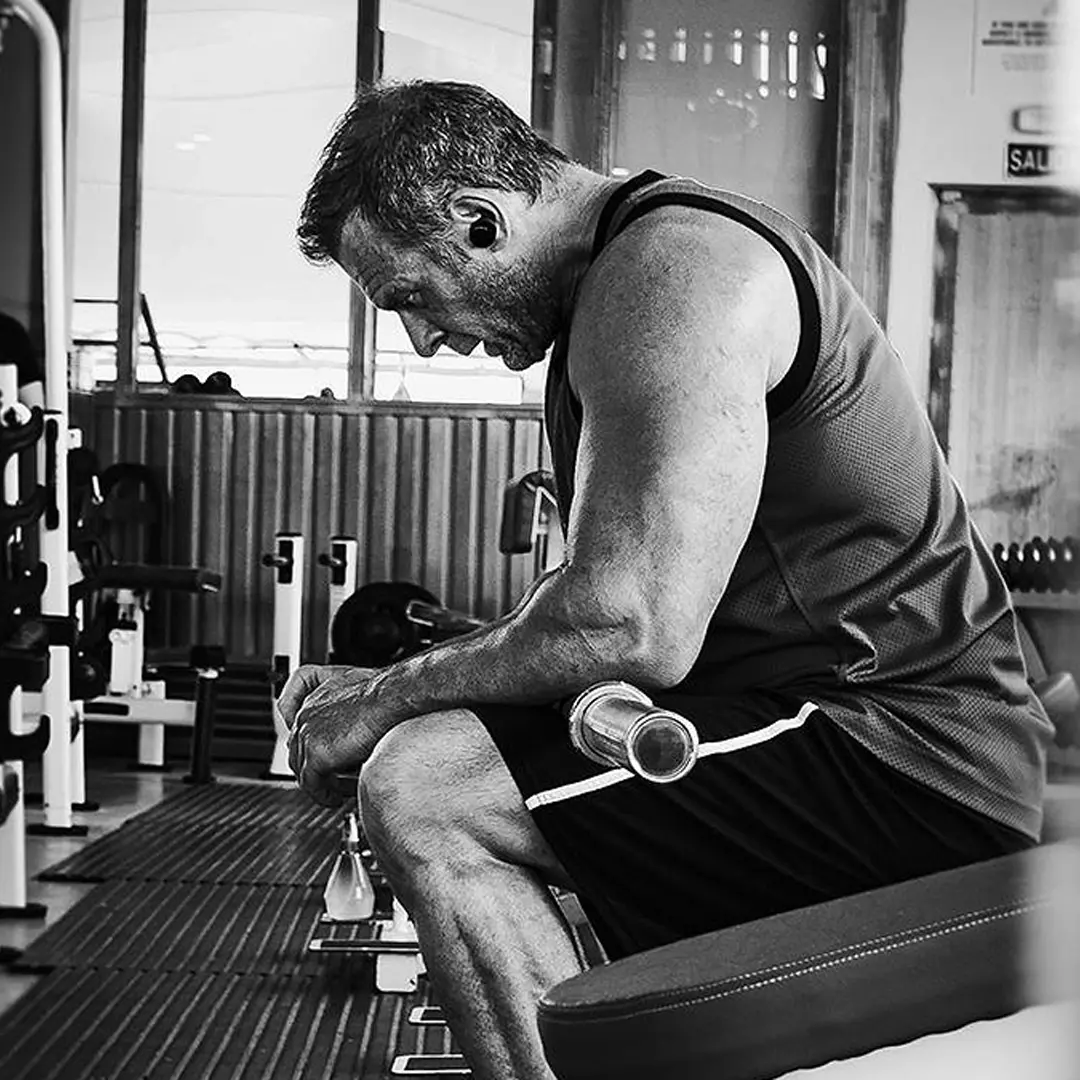 Muscular man in workout clothes sitting on a gym bench, looking down and resting with a barbell beside him.