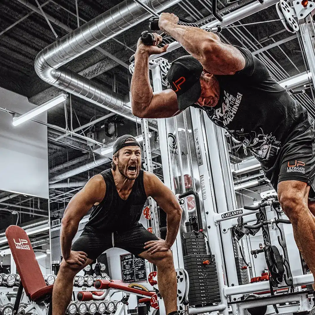 Two muscular men training intensely in a gym, one lifting weights overhead and the other encouraging him.
