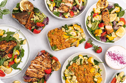 Various bowls of grilled chicken salad with mixed greens, mango, avocado, and quinoa on a light surface with two strawberries and a cup of yogurt.