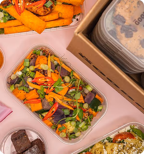 Takeout containers with colorful mixed vegetable salad, grilled polenta fries with chili slices, dark brownies, and a pasta dish on a light pink surface.