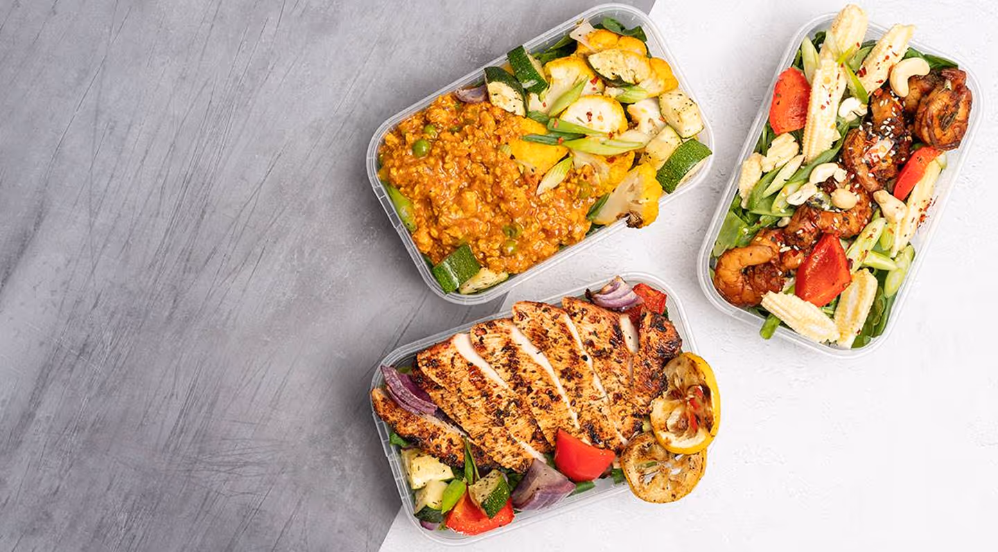 Three meal prep containers with grilled chicken, shrimp, vegetables, and a lentil stew on a two-tone gray and white surface.