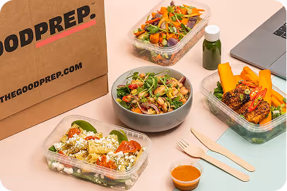 Three meal prep containers with salads and grilled chicken next to a brown paper bag and a laptop on a light surface.