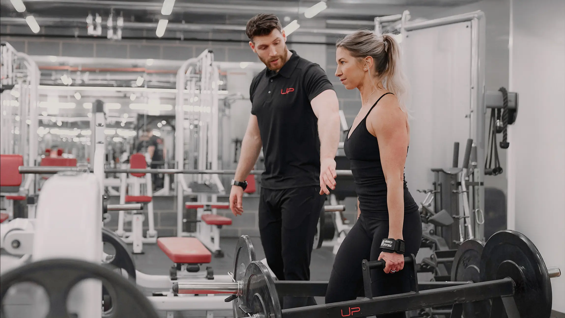 A female weightlifter preparing to lift a barbell while a male trainer instructs her in a gym.