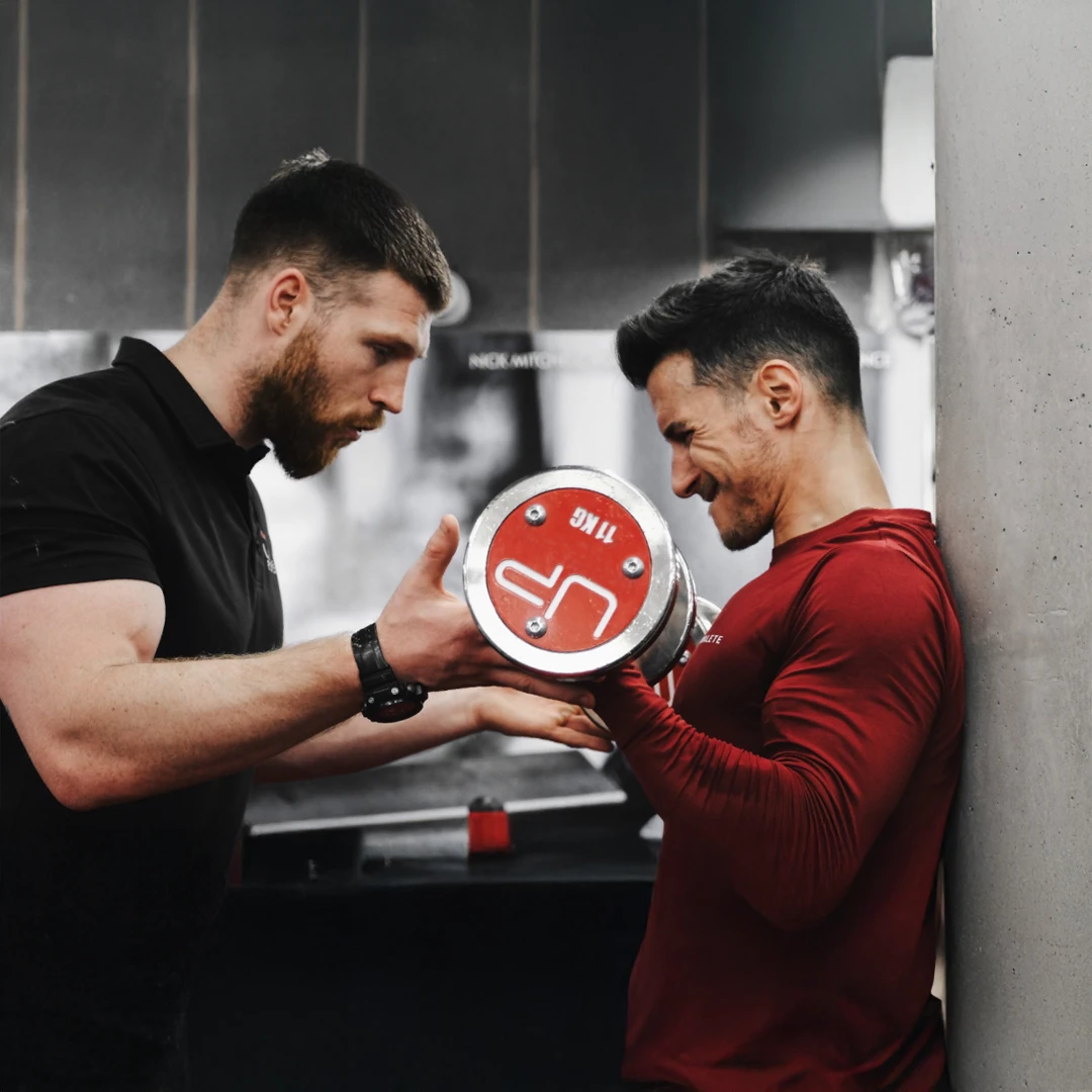 Fitness trainer closely spotting a man in a red shirt lifting a dumbbell against a wall at the gym.