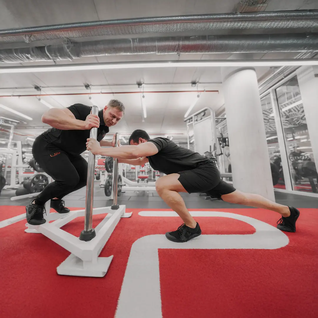 Two men in a gym engaged in a sled push exercise, one pushing and the other bracing the sled on red gym flooring.