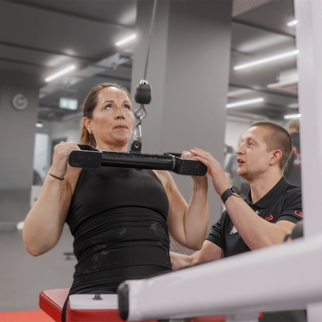 Woman doing lat pulldown exercise with trainer assisting her in a gym.