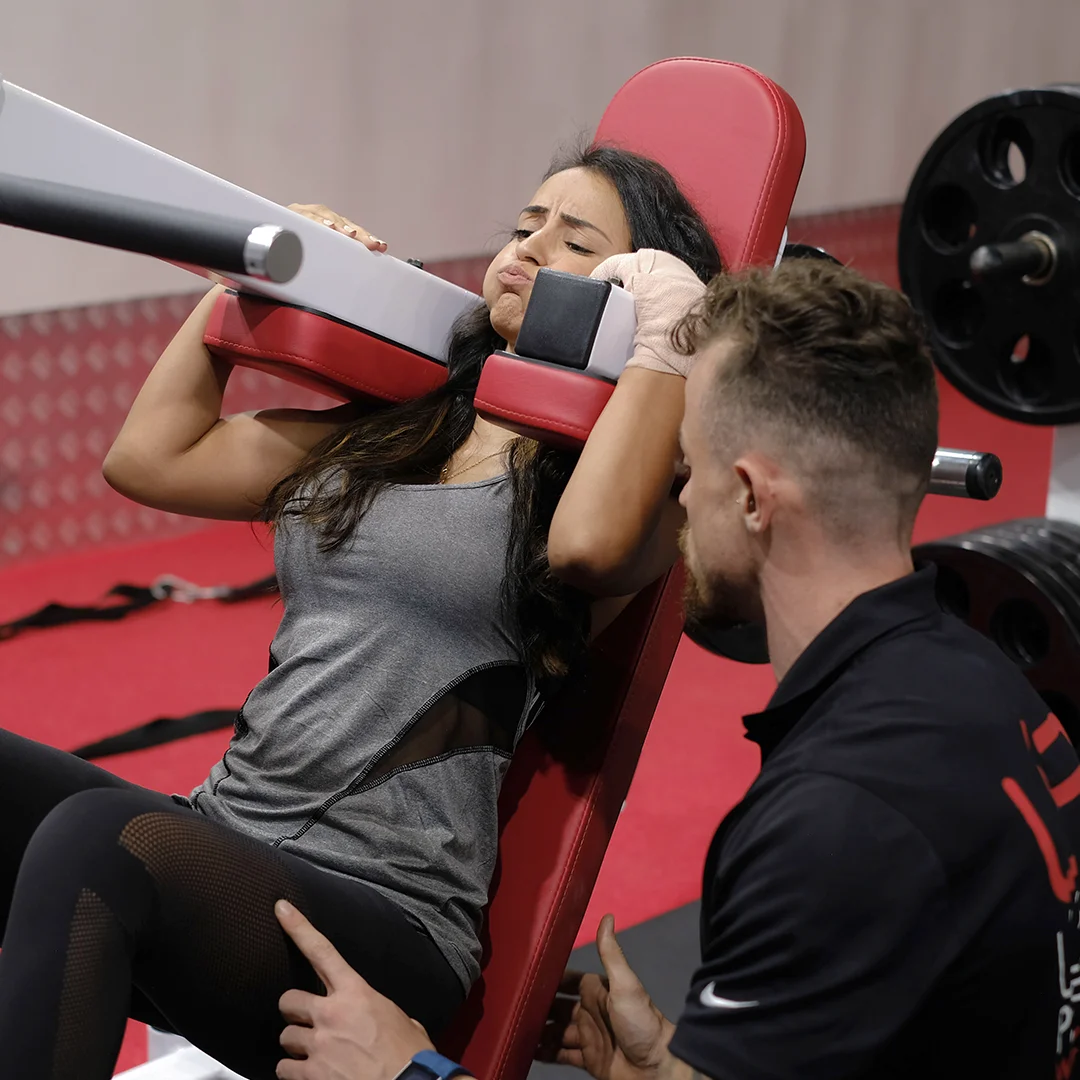 Woman in gray tank top lifting weights on a red bench with a male trainer coaching her.