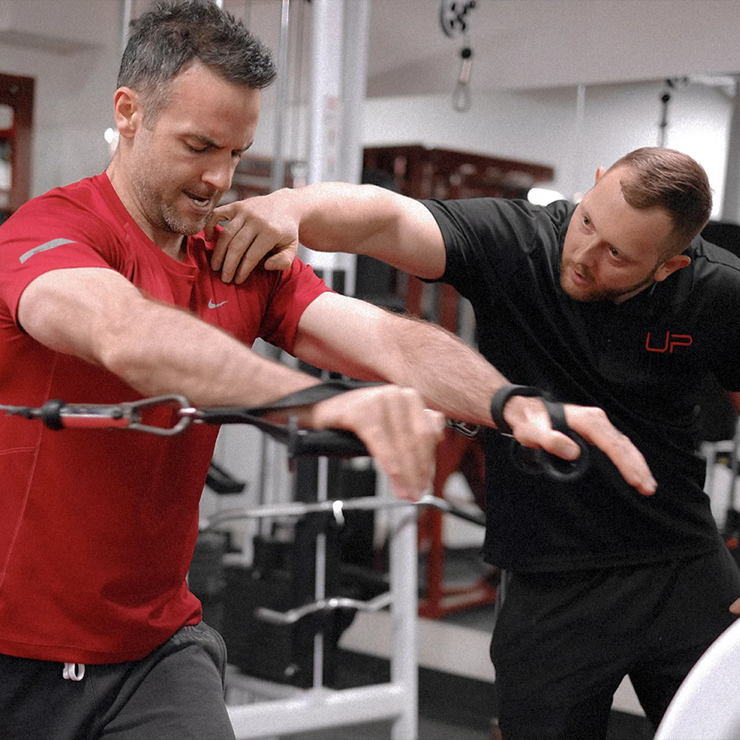 Personal trainer instructing a man in a red shirt using cable machine in a gym.