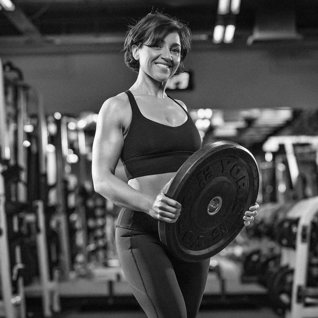 Smiling woman in workout clothes holding a large weight plate in a gym.
