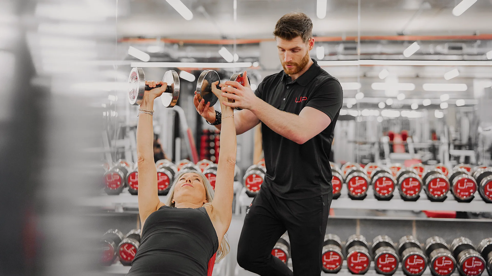 Female gym client lifting dumbbells on bench press while male trainer assists her in a gym.