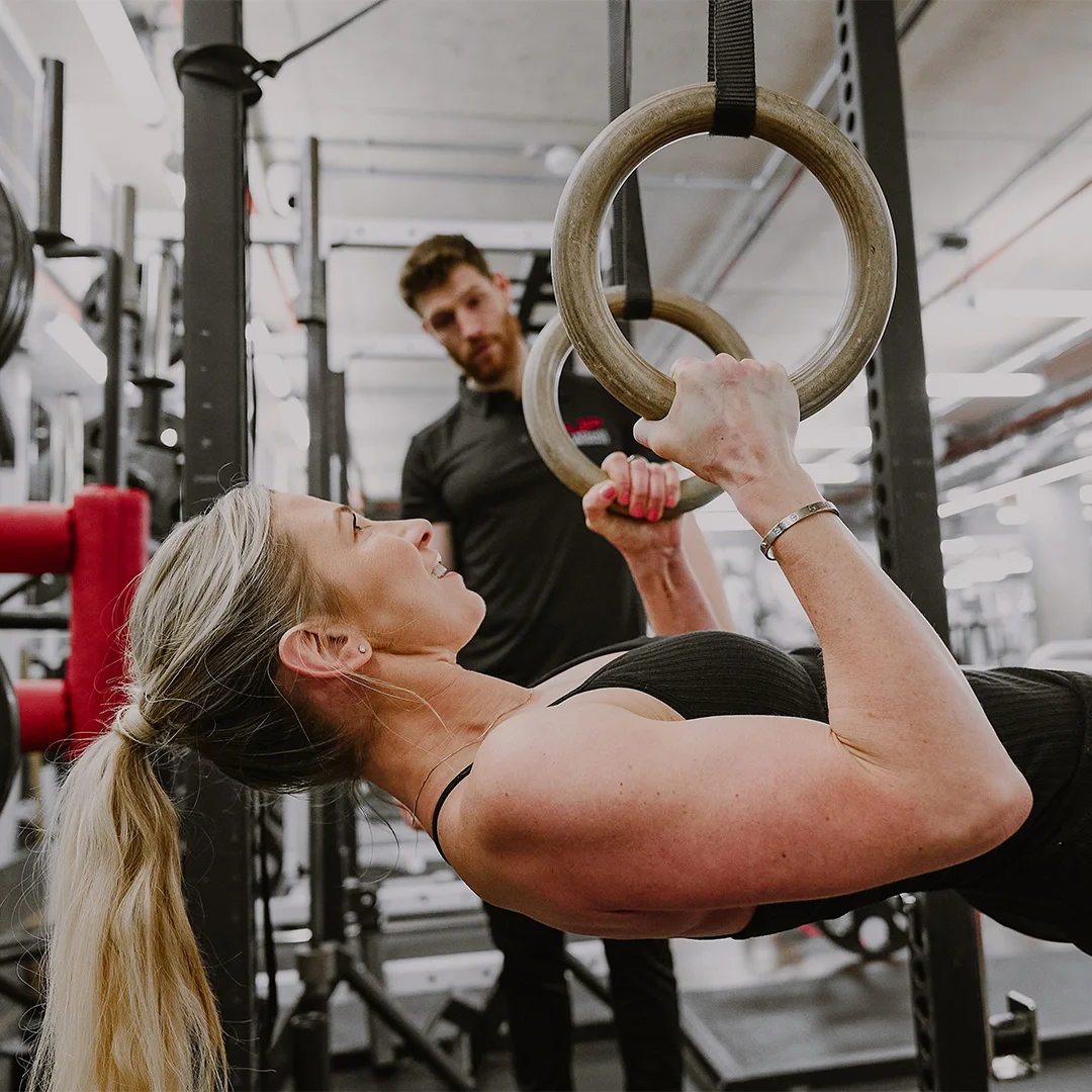 A woman doing a ring row exercise on wooden gym rings with a trainer observing her in a gym.