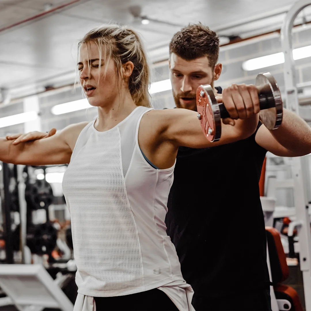 Female lifting a dumbbell sideways in a gym with a male trainer guiding her form.