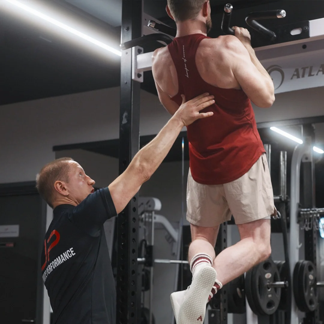 Trainer assisting a man performing pull-ups on a bar in a gym.
