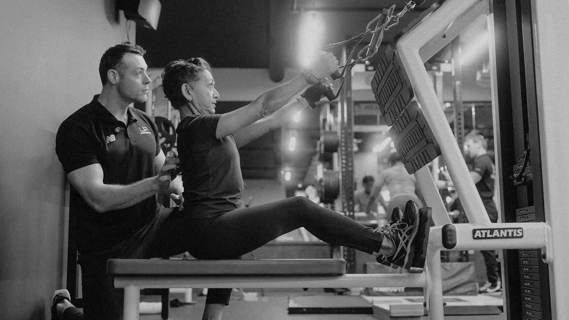 A gym trainer assists a woman using a seated rowing machine for exercise.