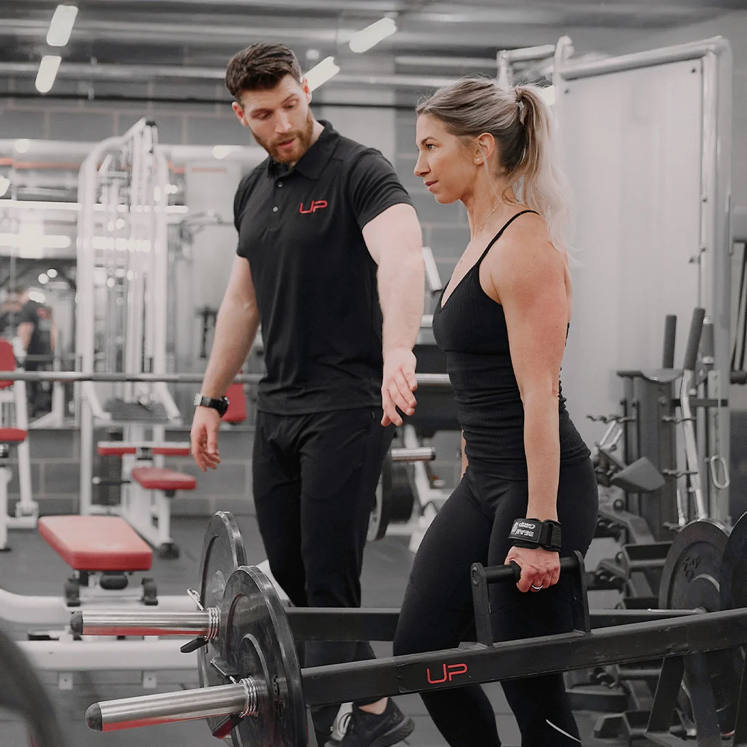 Female gym client lifting a trap bar with a personal trainer instructing her in a gym.