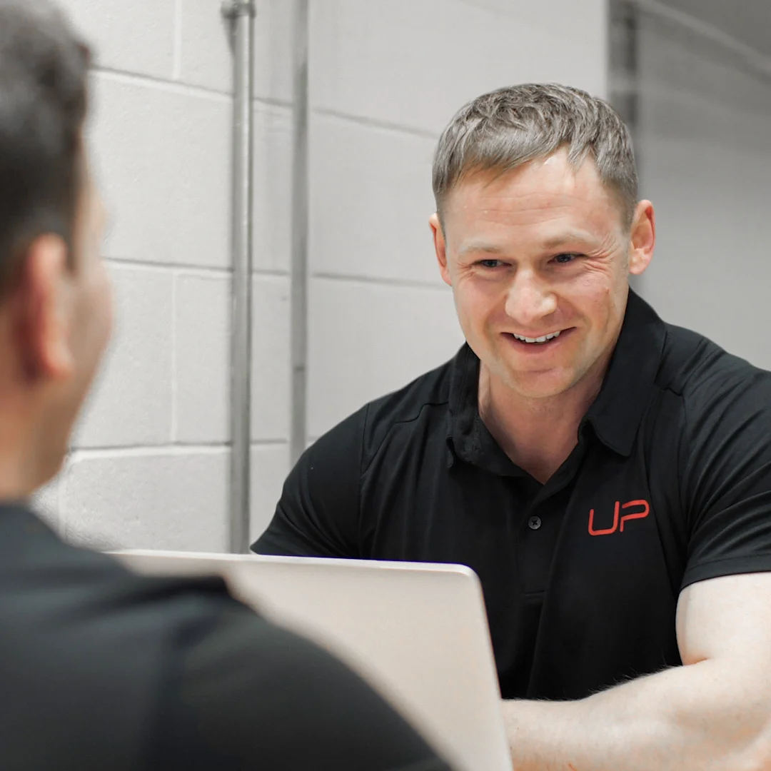 Smiling man in black polo shirt with red UP logo talking to another person over a laptop.