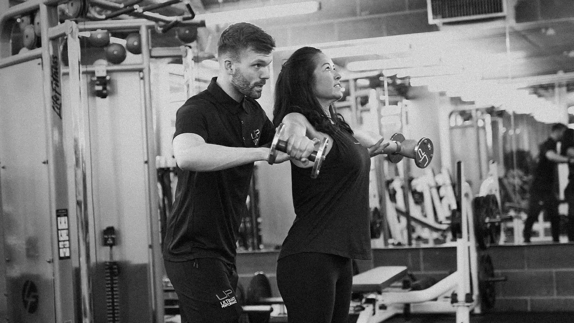 Personal trainer helps woman perform lateral raise with dumbbells in a gym.