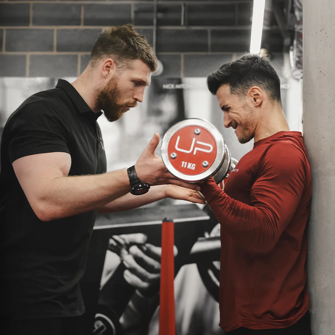 Personal trainer coaching a man lifting an 11 kg dumbbell in a gym.
