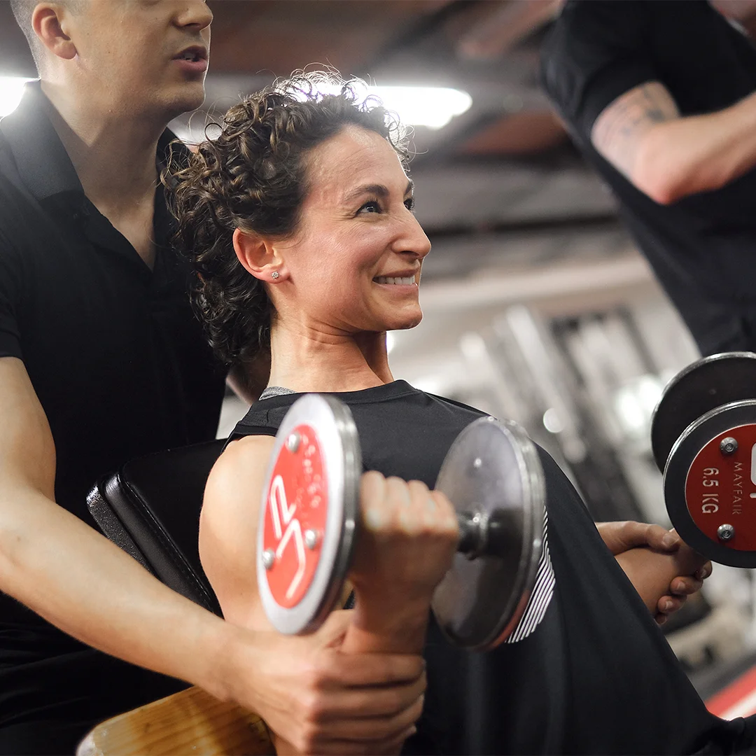 A woman smiling as she lifts dumbbells with the assistance of a personal trainer in a gym.