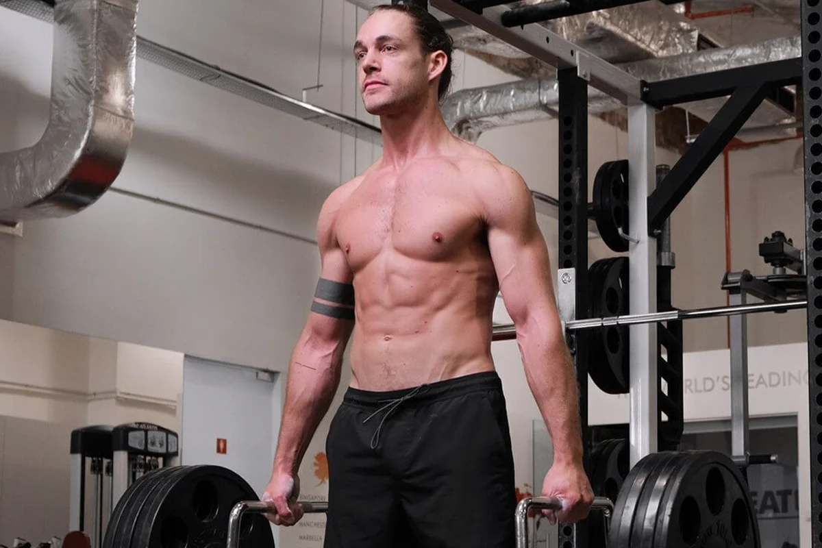 Muscular shirtless man with tattoo lifting a heavy barbell in a gym.