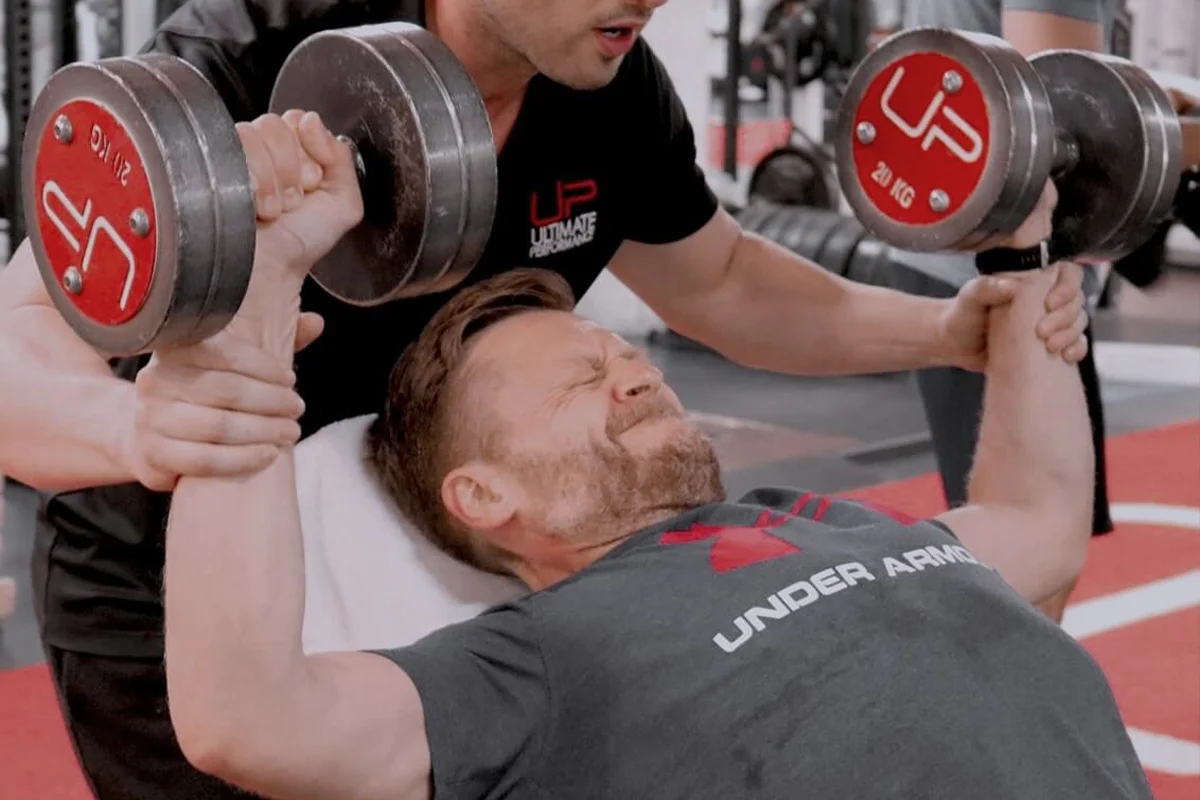 Man straining while lifting dumbbells on a bench press with a trainer spotting him in a gym.