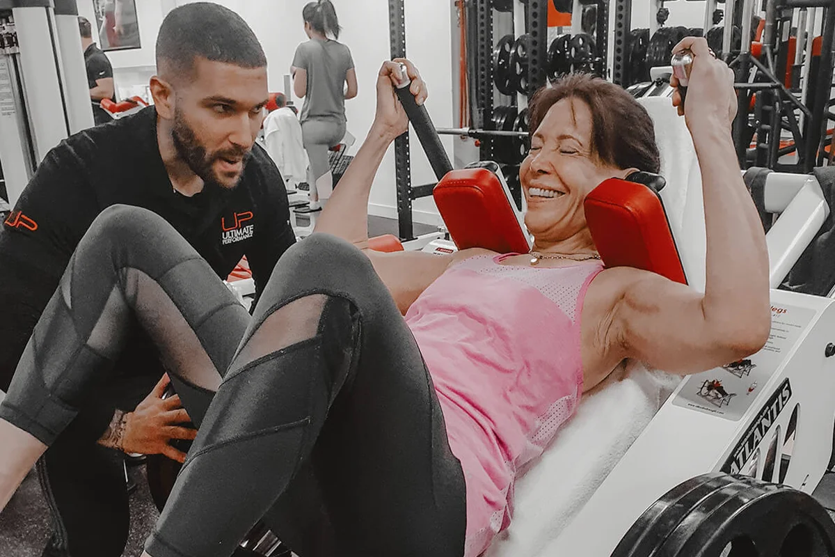 Woman in pink tank top and black leggings exercising on a leg press machine assisted by a male personal trainer in a gym.