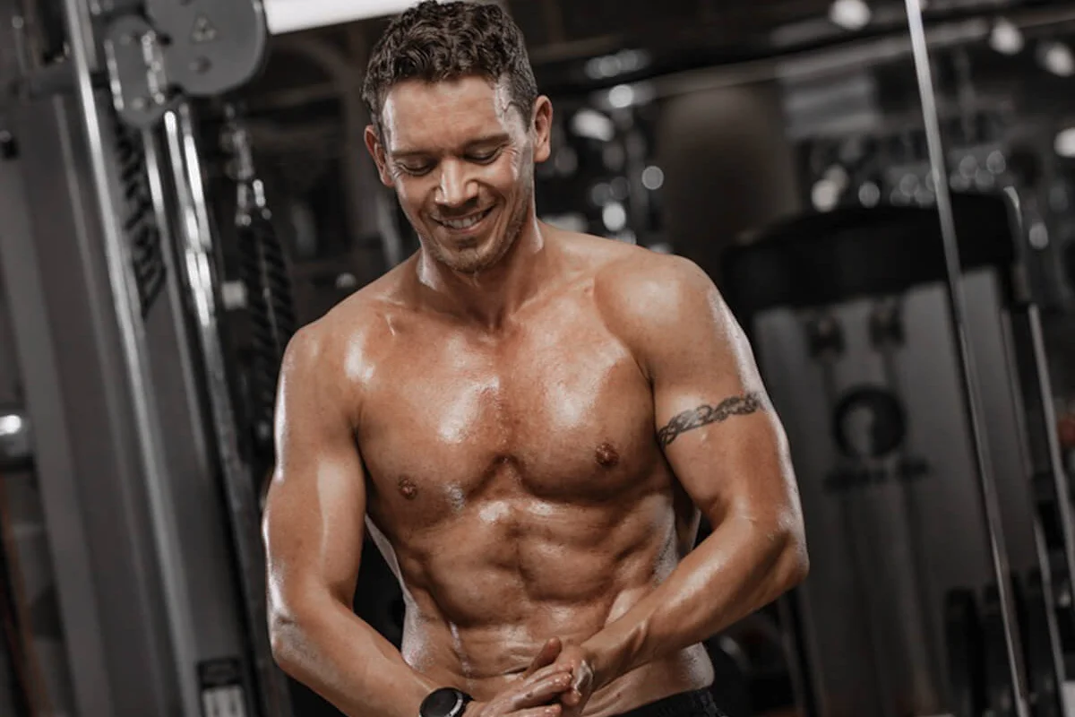 Muscular shirtless man with tattoo on upper arm smiling in a gym.