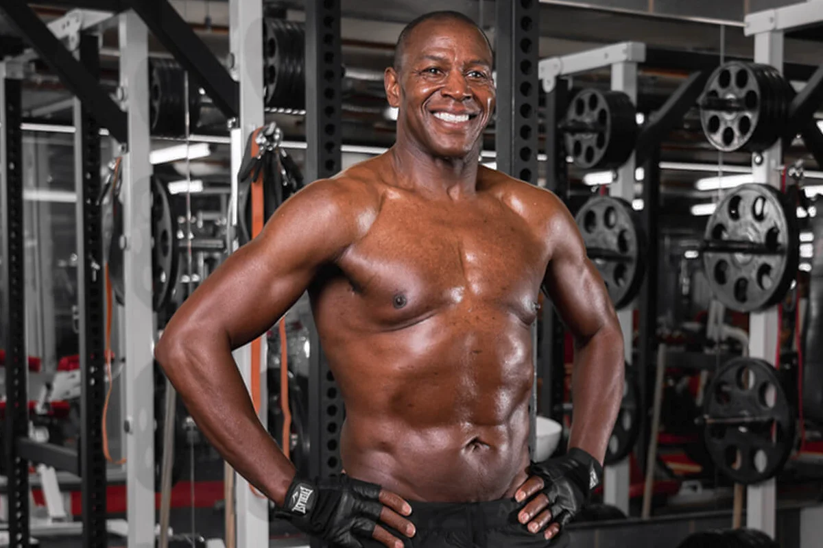 Smiling muscular man wearing black workout gloves standing with hands on hips in a gym with weight plates in the background.
