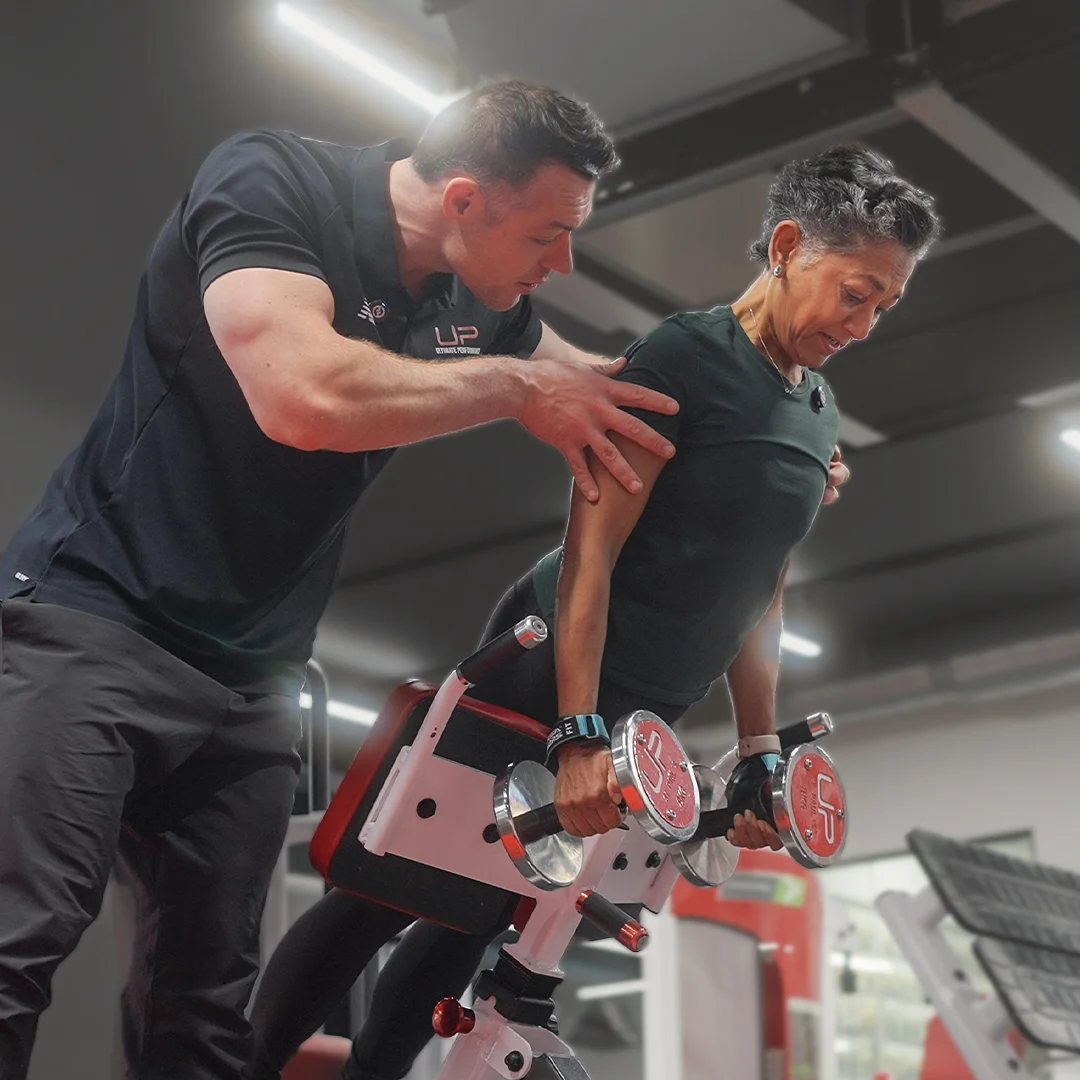 A fitness trainer assists a woman as she performs a bent-over dumbbell row exercise on a workout bench in a gym.