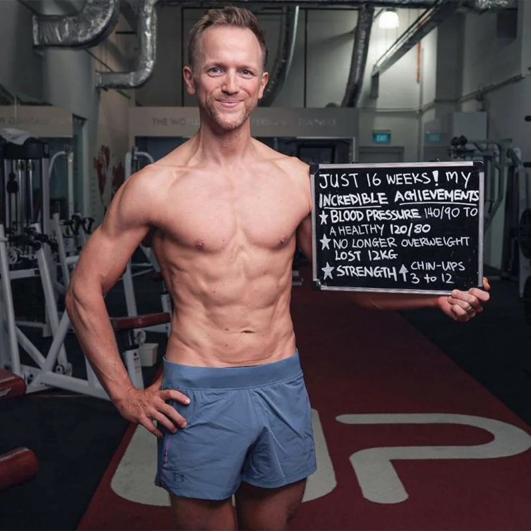 Muscular man in blue shorts holding a chalkboard listing fitness achievements including blood pressure improvement, weight loss, and increased chin-ups.